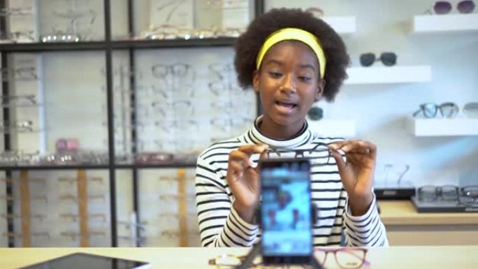 年轻女性非裔美国企业家正在时尚视频博客向手机展示眼镜，并微笑着在眼镜店在线直播销售