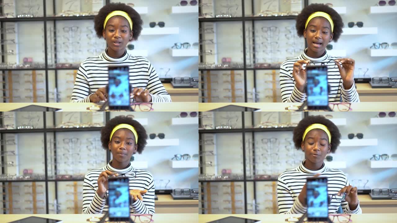年轻女性非裔美国企业家正在时尚视频博客向手机展示眼镜，并微笑着在眼镜店在线直播销售