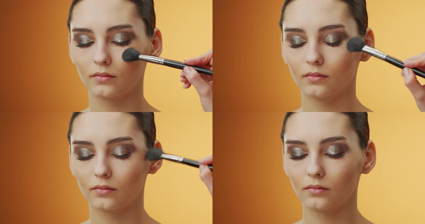 化妆申请的步骤。当化妆师用化妆刷将眼睛下方的区域除尘时，闭上眼睛的女孩脸的特写镜头。