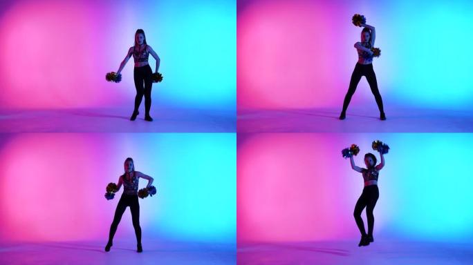 由可爱的啦啦队长在蓝色粉色工作室背景下手握绒球表演的充满活力的欢呼舞蹈。可爱的女性移动手和臀部，旋转
