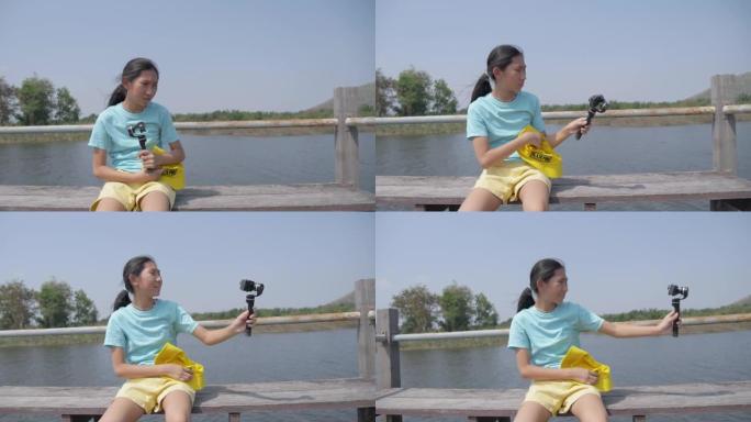 快乐的亚洲女孩自拍或在阳光明媚的日子里，在湖上的桥上用动作相机拍照，生活方式的概念。