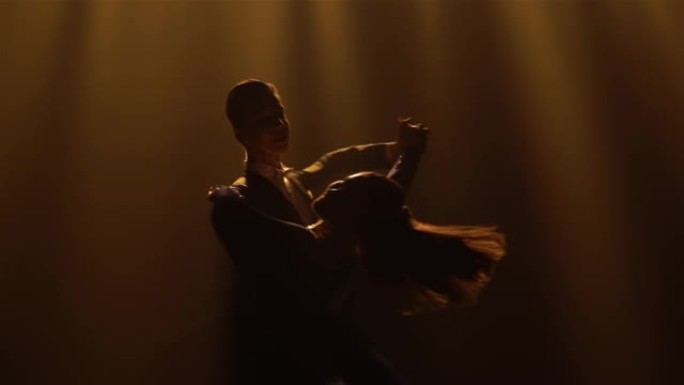 一对迷人的舞厅舞者表演的轻华尔兹动作。穿着优雅西装的男人和穿着长裙华尔兹的女人在黑暗的工作室里有黄光