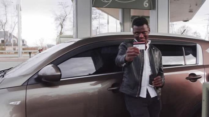 穿着便装的快乐非洲裔美国人的模糊肖像，站在自助加油站为他的现代汽车加油时，向相机展示信用卡。关注信用