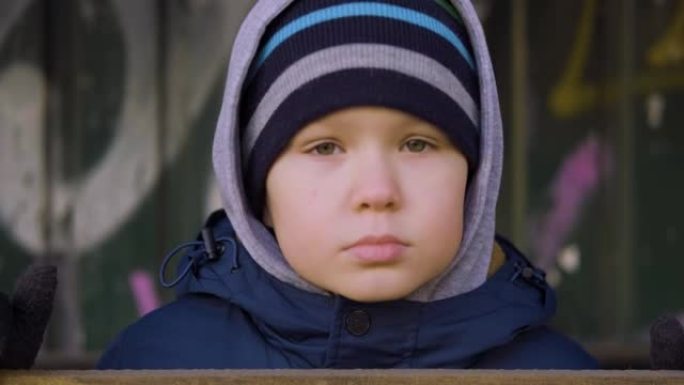 一个无家可归的小男孩紧紧抓着围栏，目不转睛地盯着镜头。流浪的孩子