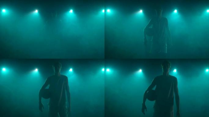 在黑暗和烟雾中，家伙的身影直接走进相机，手里拿着篮子球。篮球运动员在工作室摆姿势，背蓝灯。慢动作准备