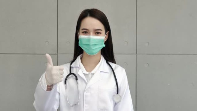 一位亚洲女医生或护士穿着制服，配有听诊器，戴着医用口罩，戴着手套，站着举起右手和拇指欣赏你。