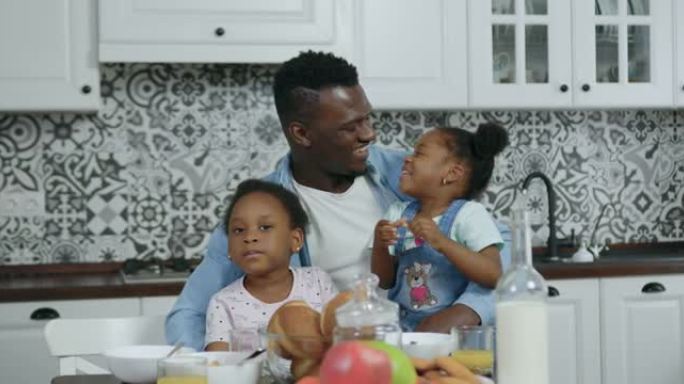 英俊快乐、充满爱心的非洲裔美国人的前视图，他们坐在厨房桌子旁，跪下抱着两个可爱的小女儿，用饼干喂养他