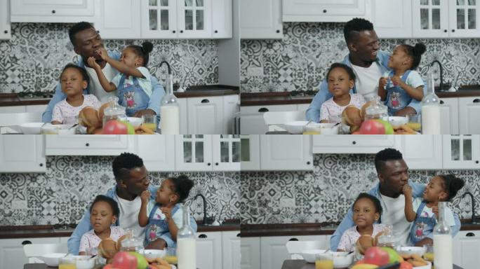 英俊快乐、充满爱心的非洲裔美国人的前视图，他们坐在厨房桌子旁，跪下抱着两个可爱的小女儿，用饼干喂养他