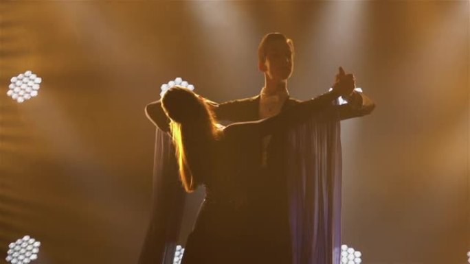 专业的舞厅舞者夫妇在明亮的灯光背景下，在黑暗的工作室里的iparket上跳华尔兹。男人和女人的舞伴在