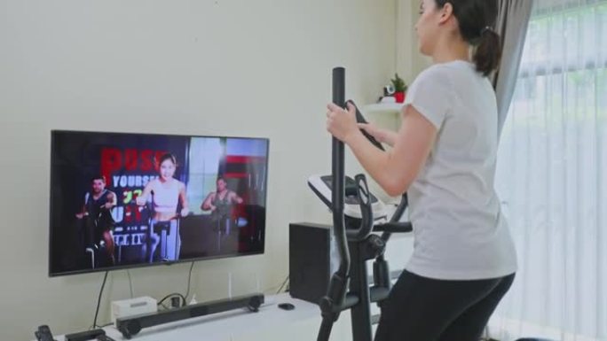 亚洲运动女性通过在家电视上观看健身现场直播或视频教程电视来锻炼。使用健身车的休闲年轻女孩在检疫过程中