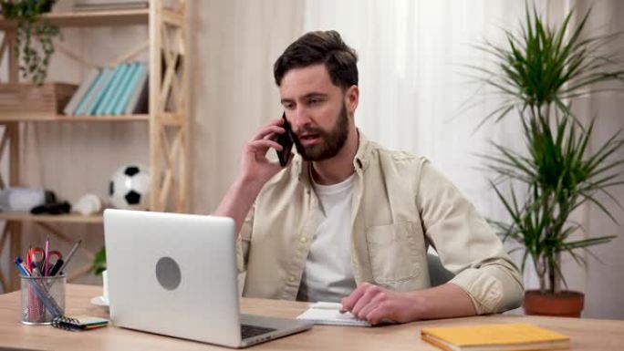 一个人坐在桌子旁，在笔记本电脑上工作，与客户或同事通电话，在笔记本上做笔记。他家庭办公室里的一个人。