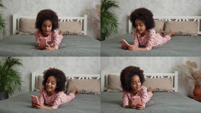 可爱的非洲裔美国小女孩看着智能手机屏幕唱歌并录制视频。穿着睡衣的少女躺在卧室的床上，室内明亮。慢动作