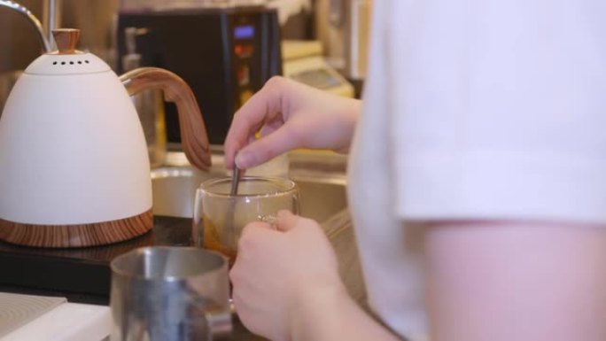 在餐厅的厨房里，服务员将白色茶壶中的水倒入带有咖啡饮料的透明杯子中，用茶匙搅拌并放下，拿起杯子并将其