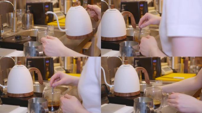 在餐厅的厨房里，服务员将白色茶壶中的水倒入带有咖啡饮料的透明杯子中，用茶匙搅拌并放下，拿起杯子并将其