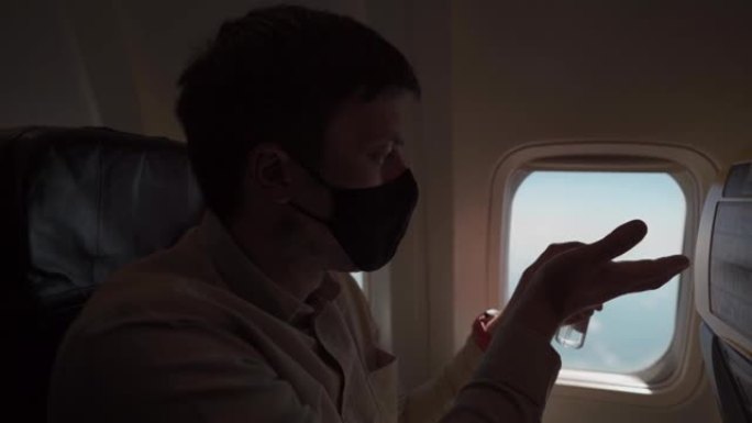 手持男子戴着面罩在飞机上使用消毒剂的镜头。带防护面罩的人使用酒精凝胶，在飞机上消毒手，新型冠状病毒肺