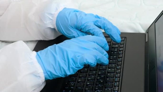 戴着防护医用手套的手在笔记本电脑键盘上打字的特写镜头。封锁期间远程在家工作