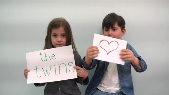 一个男孩和一个女孩展示带有铭文双胞胎和一颗心的纸，孩子们笑着玩耍，孩子们举着带有双胞胎和灰蓝色背景上
