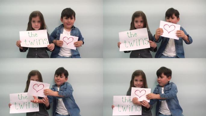 一个男孩和一个女孩展示带有铭文双胞胎和一颗心的纸，孩子们笑着玩耍，孩子们举着带有双胞胎和灰蓝色背景上