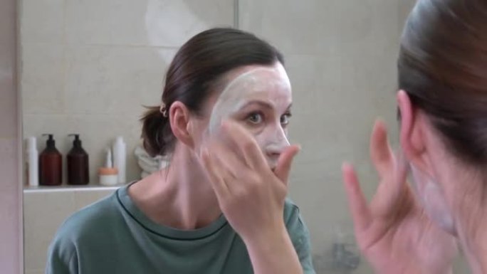 集中中年妇女在明亮的浴室近距离照镜子的脸上皮肤上涂抹保湿面膜