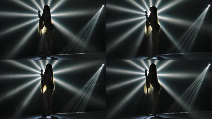 底部视图是剪影的长腿女歌手在舞台上表演的短闪亮的服装与烟雾和动态光束。歌手性感地靠近复古麦克风。慢动