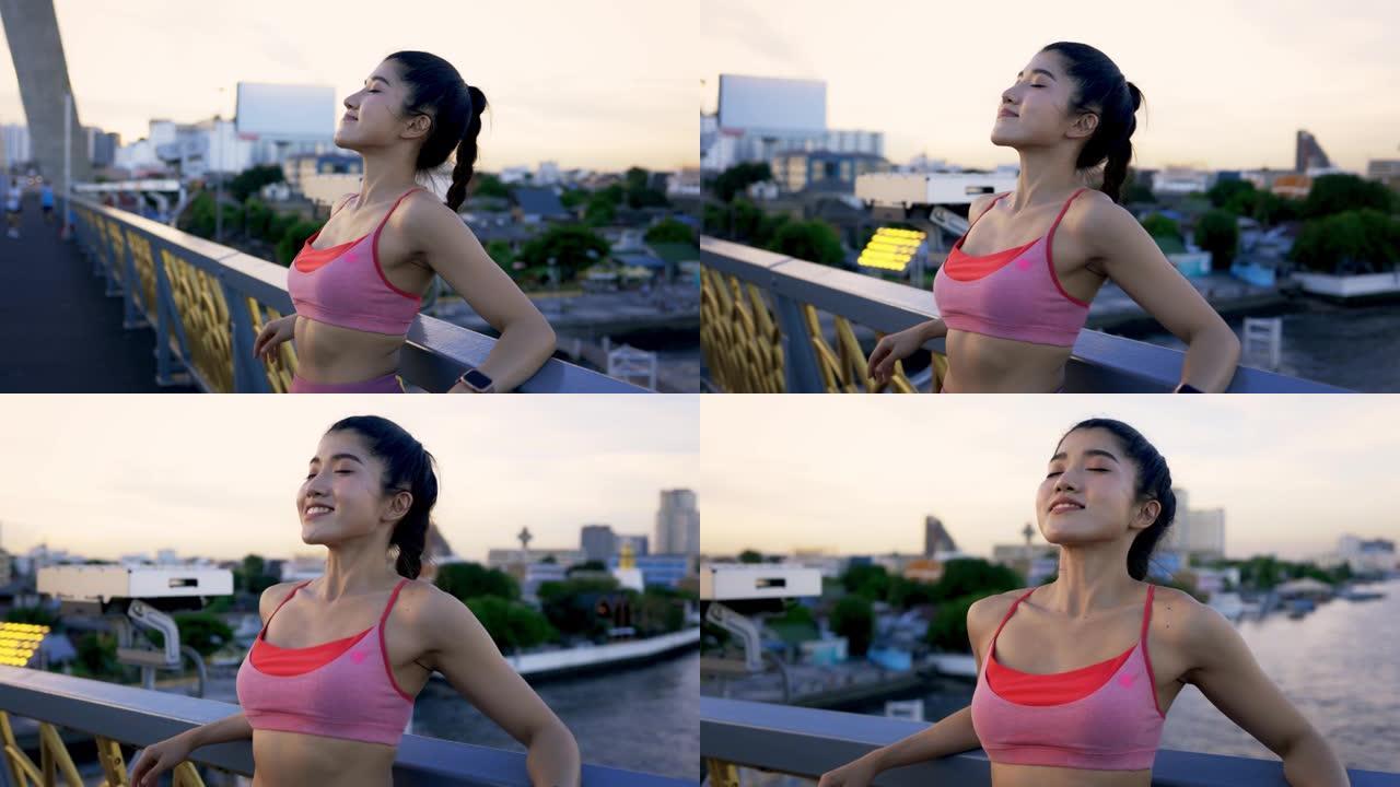 健康健康的年轻亚洲女运动员晚上跑步后靠在桥栏杆上深呼吸放松