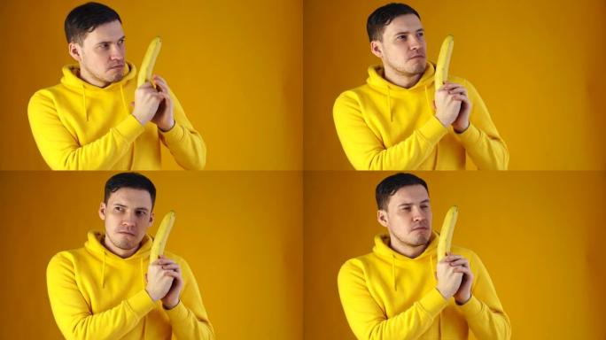 黄色背景上有香蕉的年轻人的肖像。穿着黄色连帽衫的帅哥的特写镜头玩水果，把它想象成武器