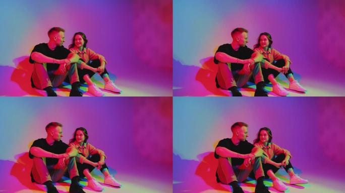 时尚快乐的年轻青少年夫妇坐在彩色霓虹灯下的地板上聊天。浪漫的约会和调情