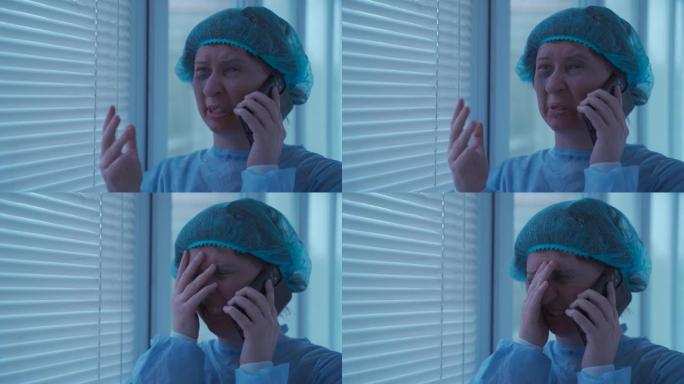 穿着一次性蓝色医用防护服和帽子的年轻女性患者不高兴，哭着打电话给急诊医院的保险公司，站在窗户旁边的走