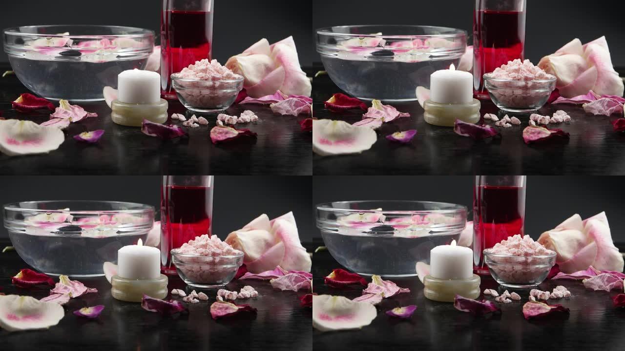 燃烧的蜡烛，香盐，玫瑰精华，玫瑰花瓣，漂浮在黑色背景上的玻璃碗中