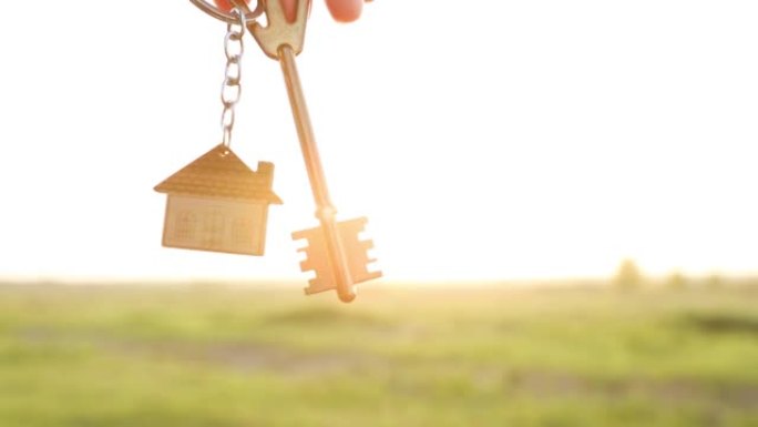 手里拿着钥匙扣的房子钥匙。天空、阳光和田野的背景。梦想家园，在农村建小屋，规划和项目设计，农场，搬到