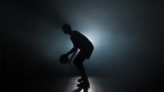 篮球运动员的剪影在黑暗的工作室里带烟和背光运球练习技巧。职业球员在篮球比赛前训练。侧视图。慢动作准备
