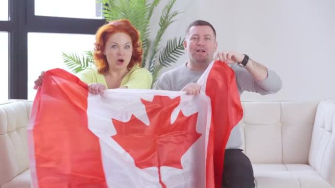快乐的一对移民坐在客厅的沙发上，手持加拿大国旗唱国歌。快乐快乐的男人和女人享受移民到加拿大的视频聊天