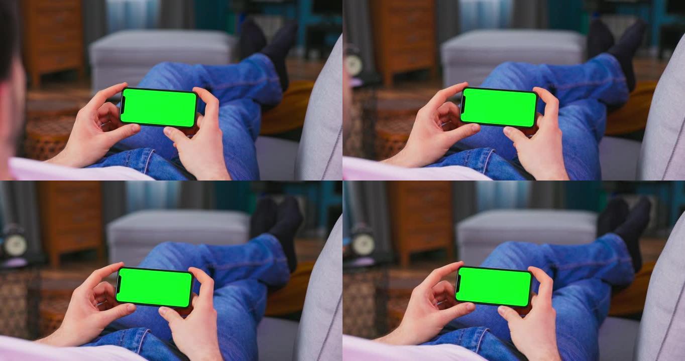 在家中的年轻人在水平景观模式下使用绿色模拟屏幕手机在沙发上休息。男孩正在浏览互联网，观看视频，社交媒