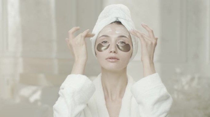 年轻女子在镜子里看着自己，穿着浴袍，用毛巾用手在脸上涂上保湿霜。