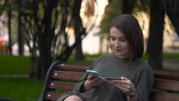 拿着信用卡和电话的学生在街上，黑发女人坐在长凳上，通过智能手机上的银行卡通过互联网进行在线支付