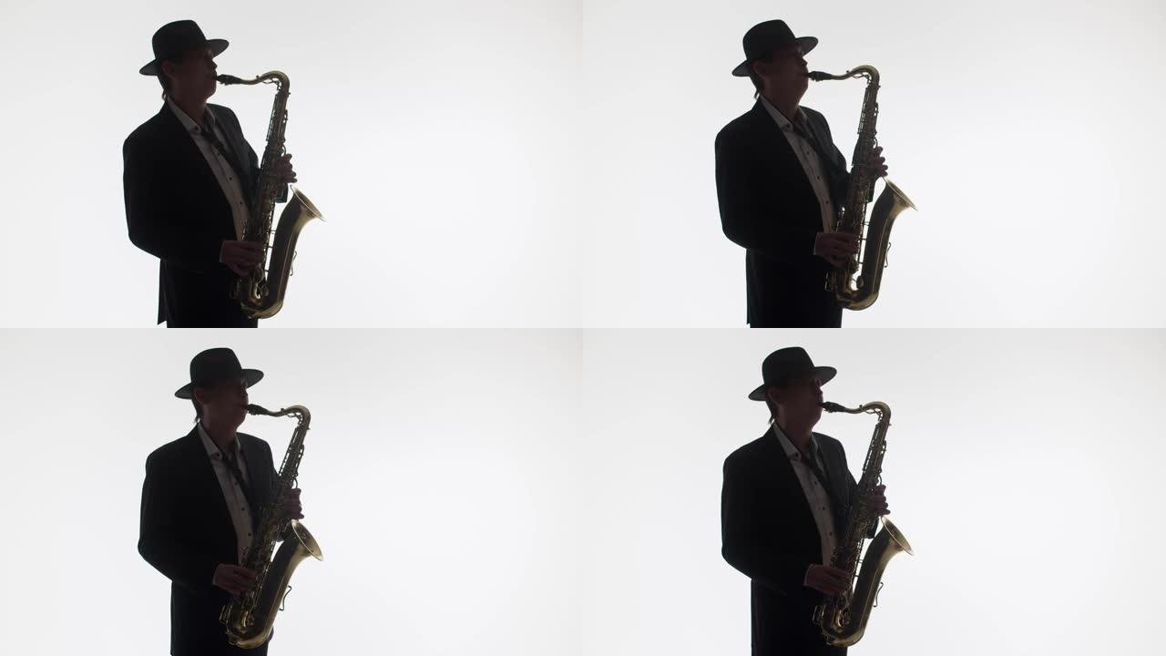 一位优雅的音乐家在工作室的白色背景上演奏金色萨克斯管的深色剪影。穿着帽子和西装的艺术家演奏萨克斯管。