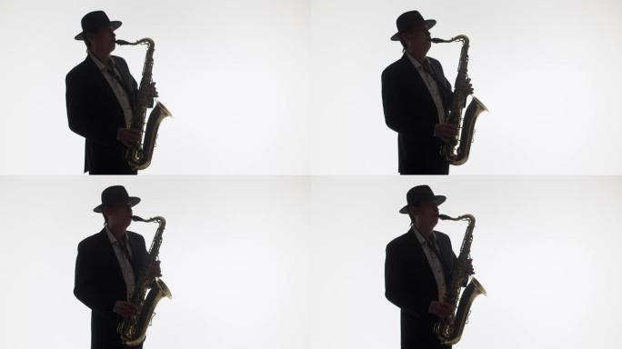 一位优雅的音乐家在工作室的白色背景上演奏金色萨克斯管的深色剪影。穿着帽子和西装的艺术家演奏萨克斯管。