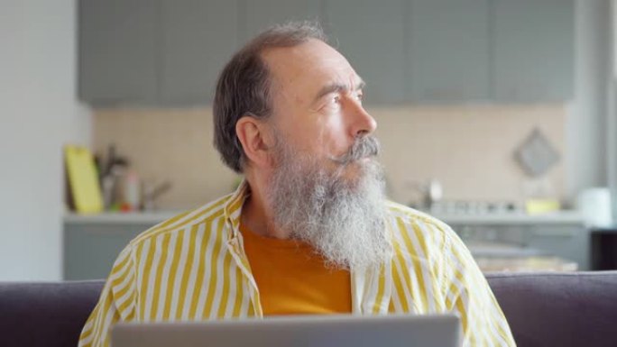 在冠状病毒大流行期间，坐在沙发上、在家工作的长白胡子的老人在笔记本电脑上打字，思考和微笑