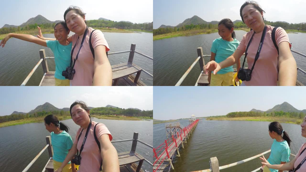 快乐的亚洲母亲和她的女儿在阳光明媚的日子里一起在湖上的桥上自拍，旅行生活方式的概念。
