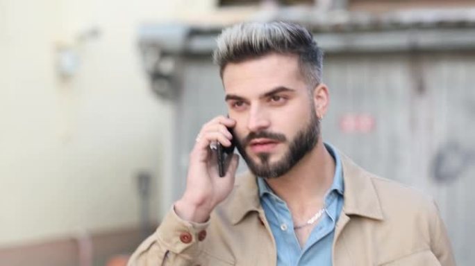 穿着米色夹克的酷未刮胡子的男人在电话里聊天，自信地走在外面的灰色车库旁
