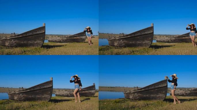 在炎热的晴天，穿着草帽和牛仔短裤的迷人年轻女子在岸上的废弃木船附近行走。跟踪拍摄4k 50 fps慢
