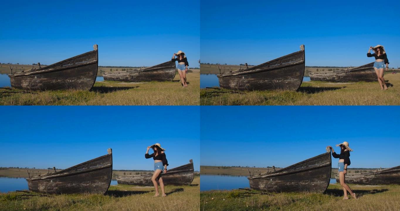 在炎热的晴天，穿着草帽和牛仔短裤的迷人年轻女子在岸上的废弃木船附近行走。跟踪拍摄4k 50 fps慢