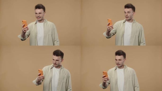 时尚男人的肖像正在使用智能手机进行视频通话。穿着米色衬衫的男性时装模特在棕色工作室背景下摆姿势。特写