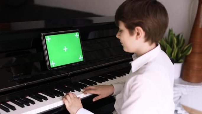 学校男孩学习弹钢琴与平板电脑应用程序。孩子喜欢学习演奏乐器。平板电脑与绿屏色度键背景新的应用程序，商