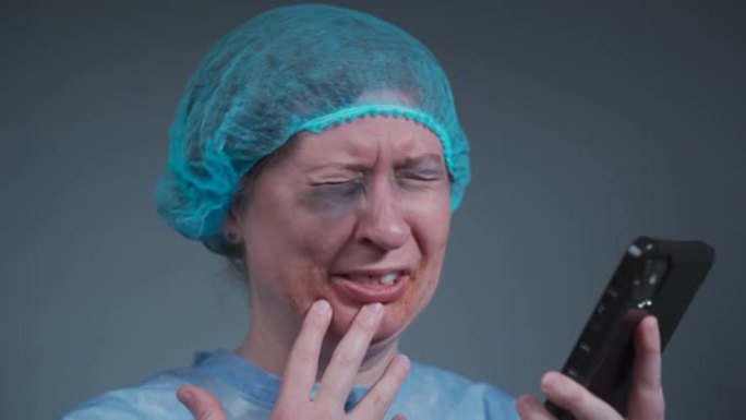穿着一次性蓝色医用防护服的女性患者哭泣并打电话给急诊医院的保险公司。在诊所发生暴力事件后面部受伤的女