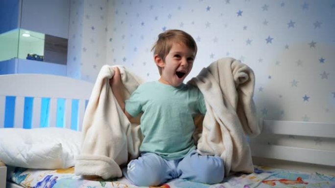 穿着睡衣的滑稽笑的蹒跚学步的男孩的慢动作晚上躲在毯子下跳出来。吓唬和吓唬孩子。