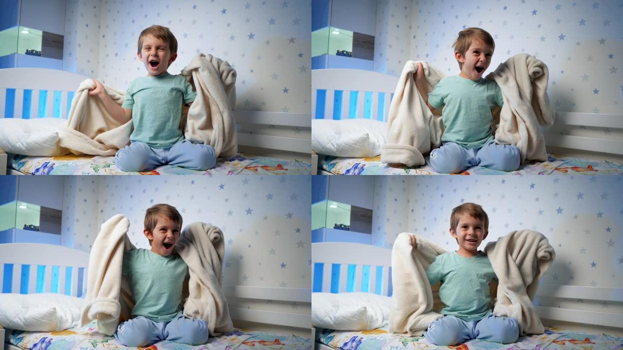 穿着睡衣的滑稽笑的蹒跚学步的男孩的慢动作晚上躲在毯子下跳出来。吓唬和吓唬孩子。