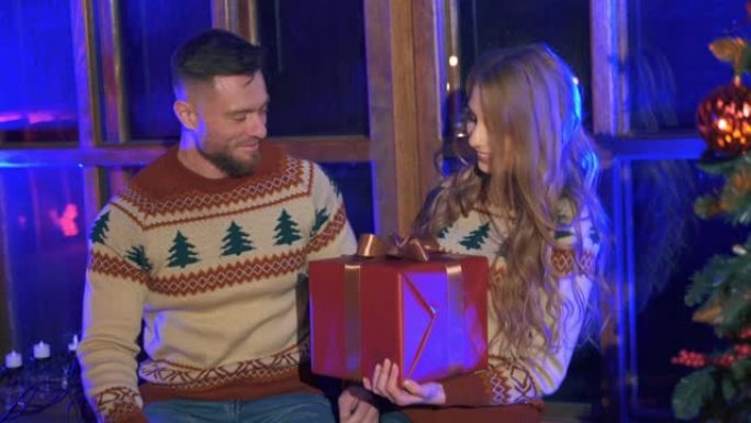 可爱的夫妇穿着圣诞毛衣。英俊的男人给心爱的女孩一个带礼物的红色盒子。快乐女人收到男友送的新年礼物。