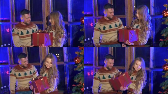 可爱的夫妇穿着圣诞毛衣。英俊的男人给心爱的女孩一个带礼物的红色盒子。快乐女人收到男友送的新年礼物。