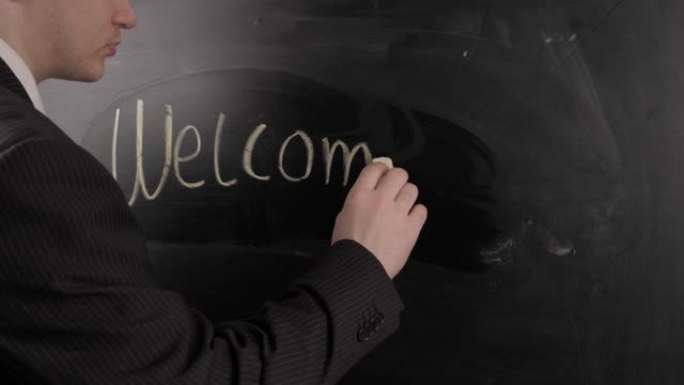 穿着黑色西装的老师在黑板上写下了 “欢迎” 一词。